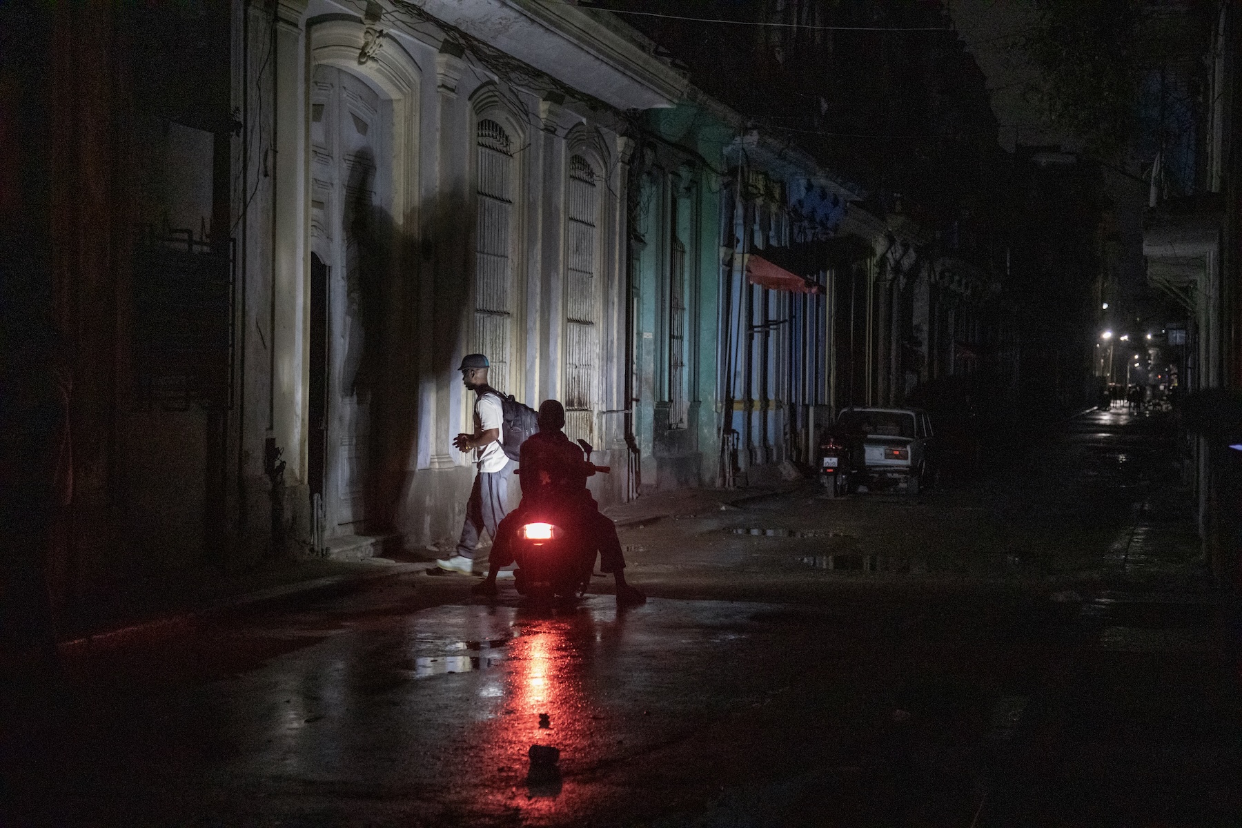 Photo of a person riding a scooter on a dark road at night. The scooter's headlights cast the shadows of nearby people on the side of the stone building.