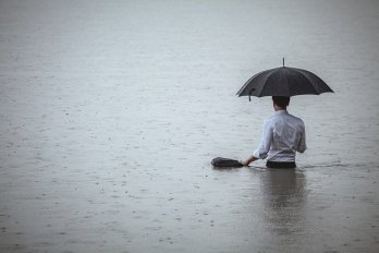 Rear view of a man wearing white shirt and holding an umbrella and a briefcase, standing waist-deep in the water on a rainy day