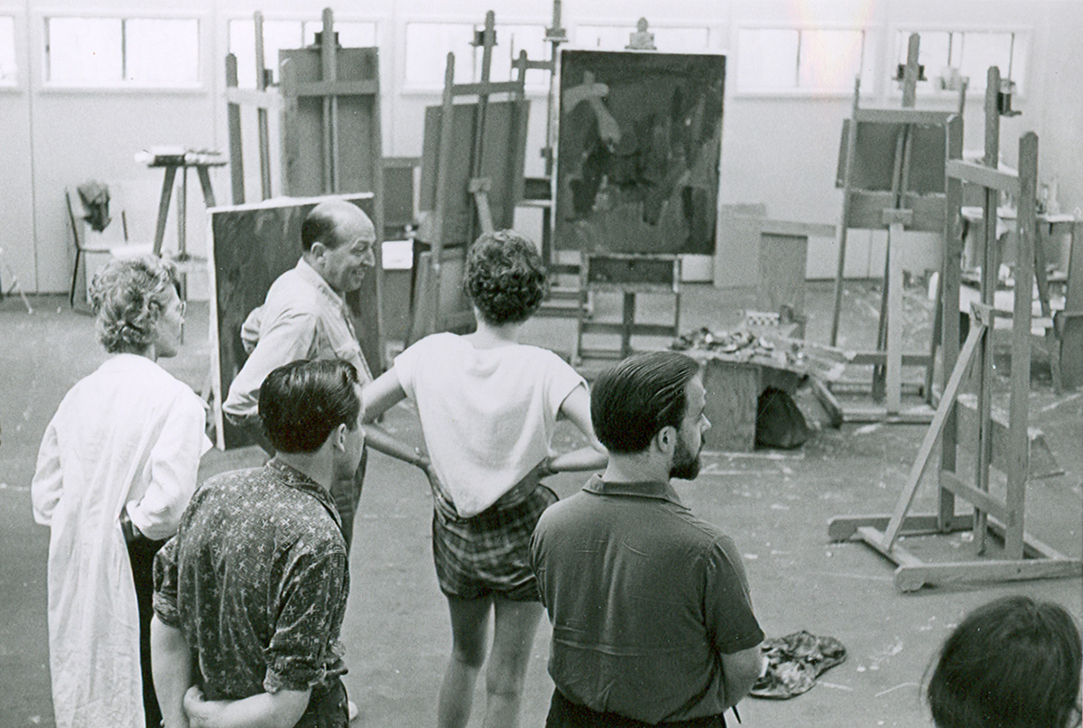Black and white photo of Clement Greenberg, an older balding man, talking with a group of students. In the background are numerous easels and abstract paintings.