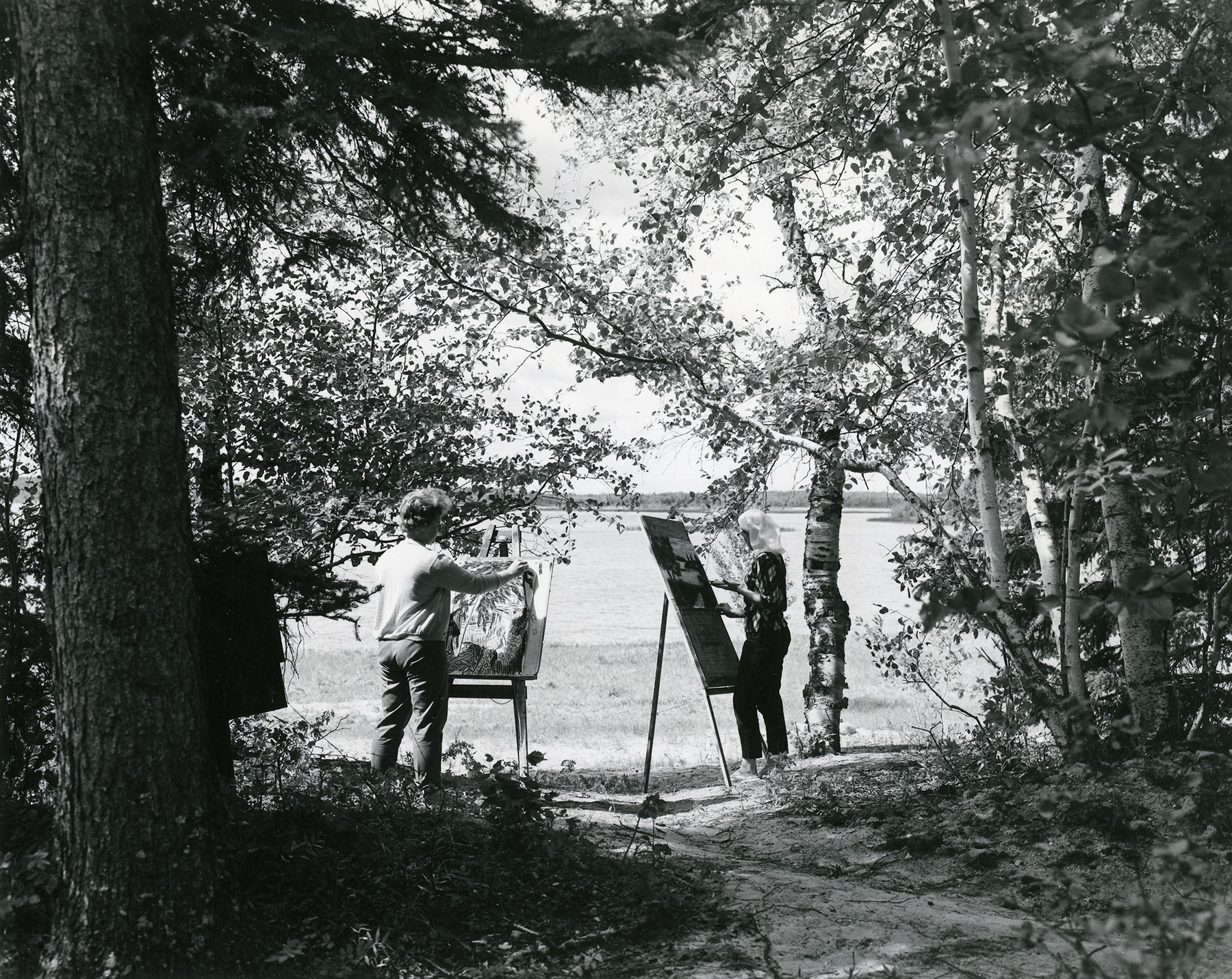 Black and white photograph of two women, surrounded by trees, standing in front of easels and painting. A shoreline and lake is visible in the distance.