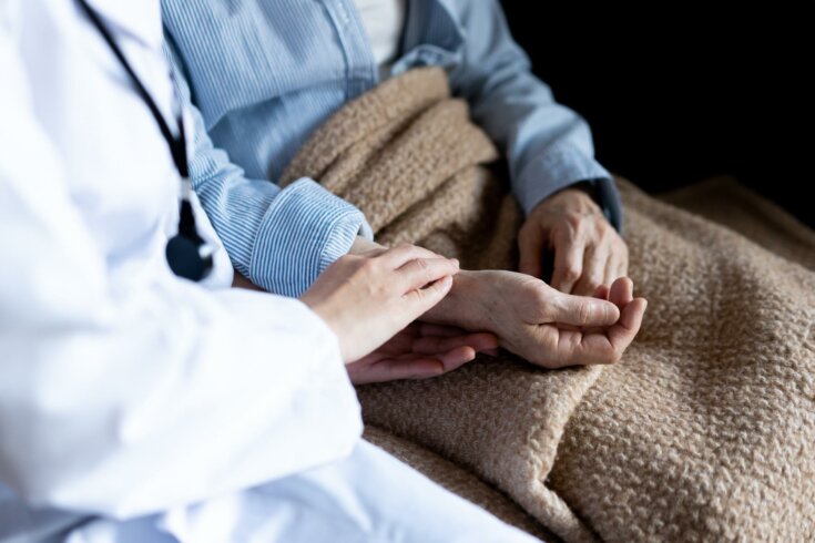 Photo of a doctor in a white coat holding hands of another person in blue pajamas with a brown blanket over their lap.