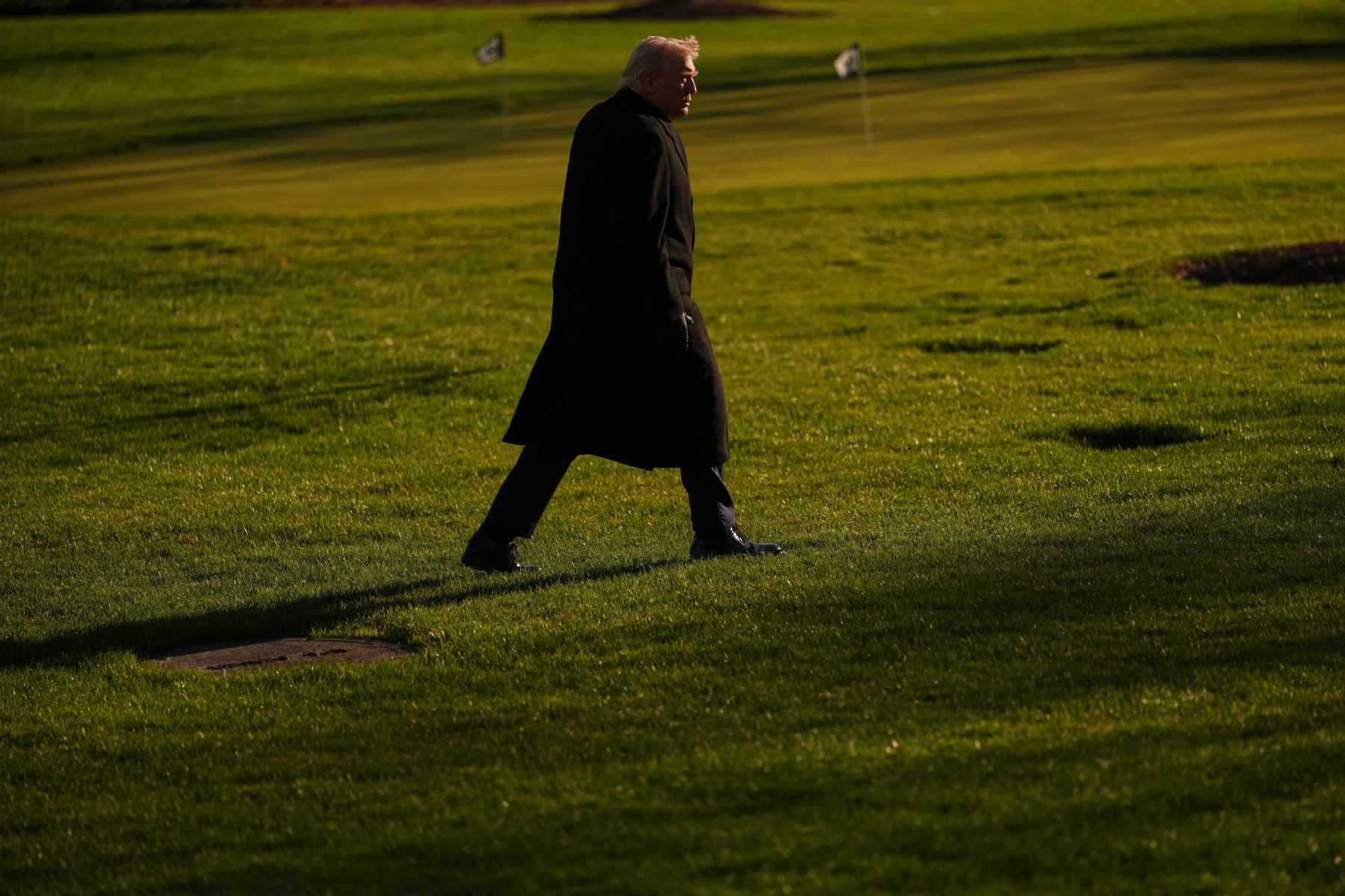 Photo of President Donald Trump walking across green grass while wearing a long, black coat.