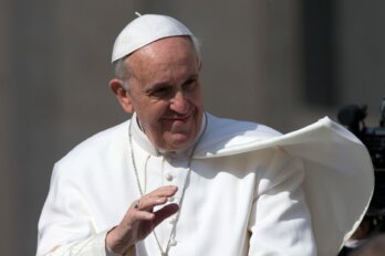 A photo of Pope Francis dressed in white, his hand partially raised as his cape flutters in the wind
