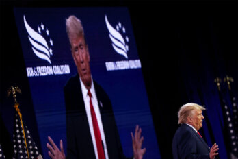 A photo of Donald Trump speaking at a conference on June 2023 while a projector displays an enlarged real-time broadcast of him in the background.