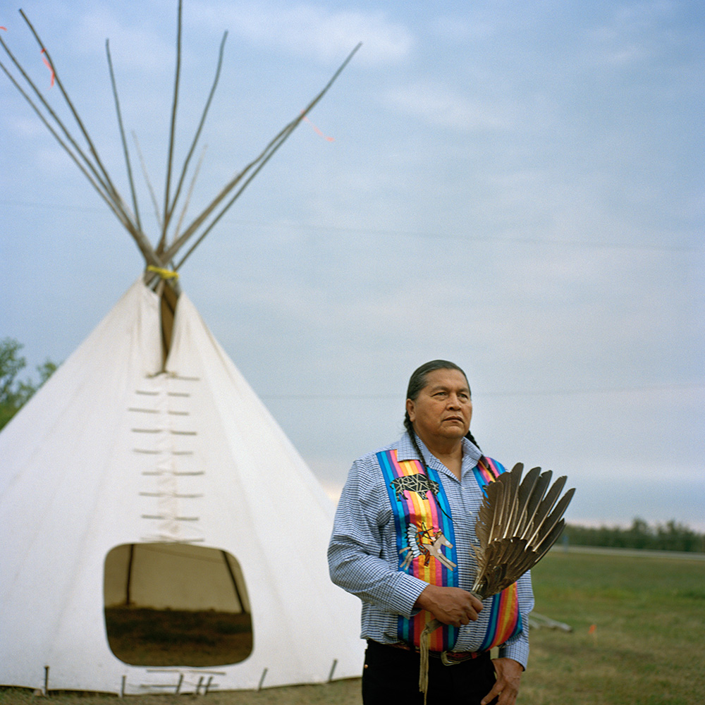 Photograph of Alvin Baptiste. He is holding a traditional fan made of brown-and-white eagle feathers and standing in front of a tipi.