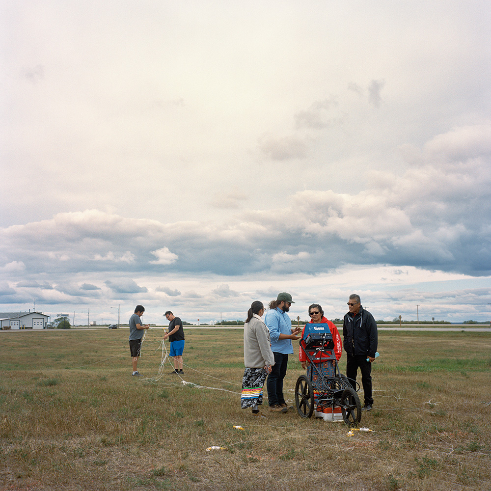 Photograph of a group of elders, community members, and SNC-Lavalin staff standing in a field beside a highway. Four people in front surround a cart holding ground-penetrating radar equipment. Two others begin to lay out a grid of survey lines with string.