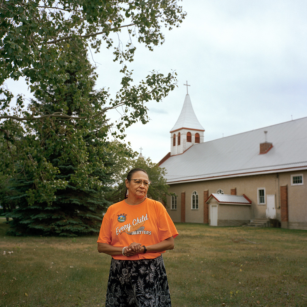 Photograph of Karen Whitecalf. She is standing in front of a church and wearing an orange t-shirt that reads “Every Child Matters.”