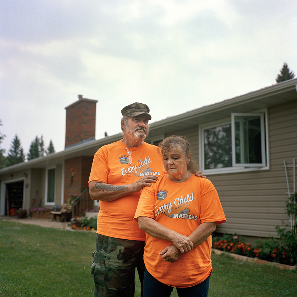 Photograph of Doug Montgomery and Donna McBain standing in front of their house. They are wearing orange t-shirts that read “Every Child Matters.”