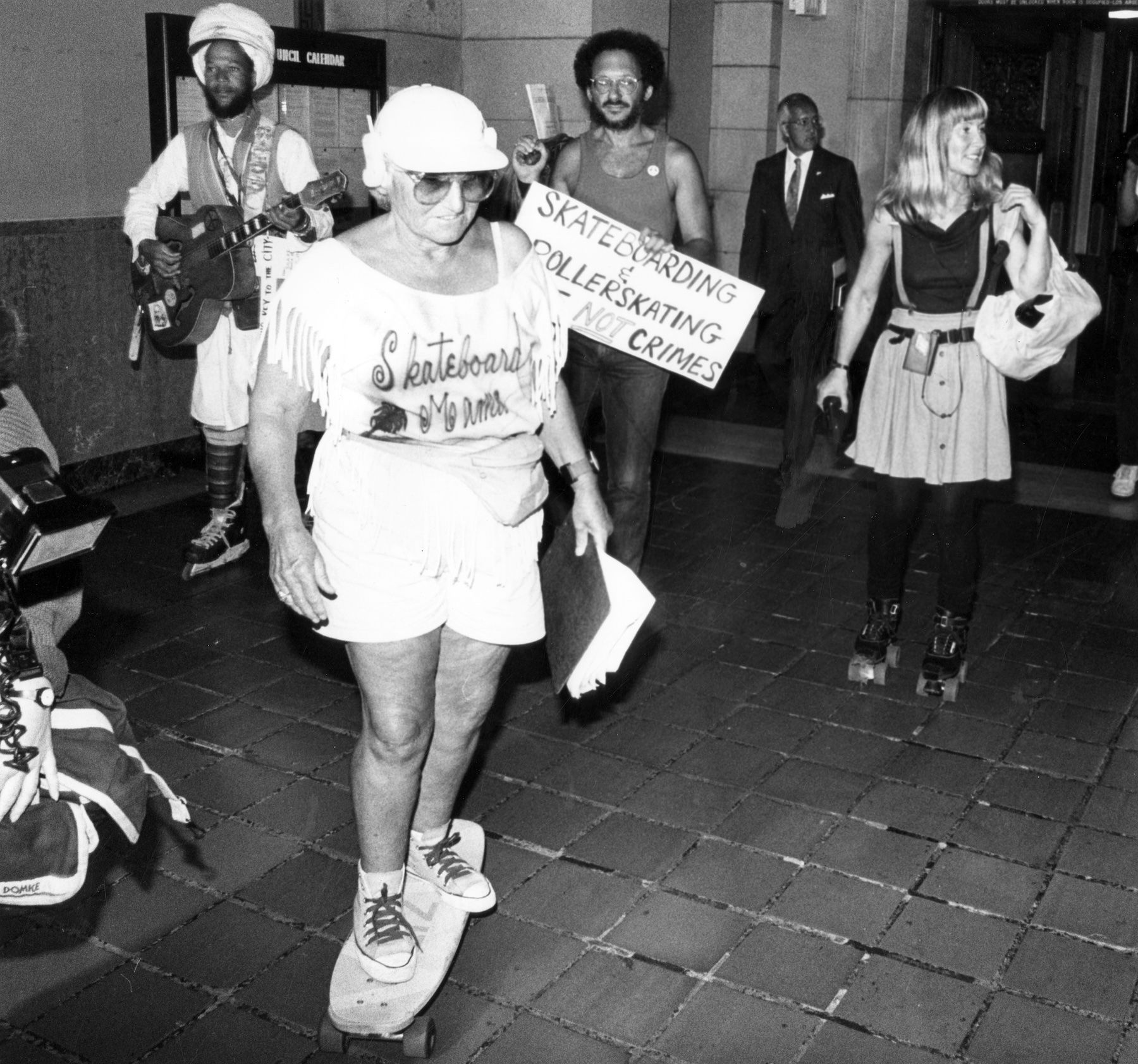Liz Bevington is a senior citizen riding her skateboard on a cobbled street. She is wearing a fringed T-shirt that says ‘Skateboard Mama,’ white shorts, a hat, and sunglasses. Liz is part of a protest, and there are at least three other people rollerskating in the photo.