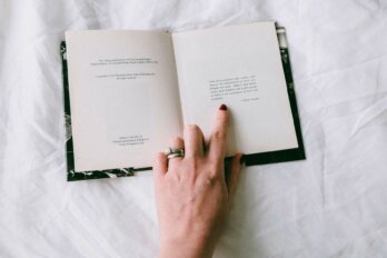 A book lies open to the opening epigraph. A finger with dark red nail polish, points to the quote, the hand resting on the book