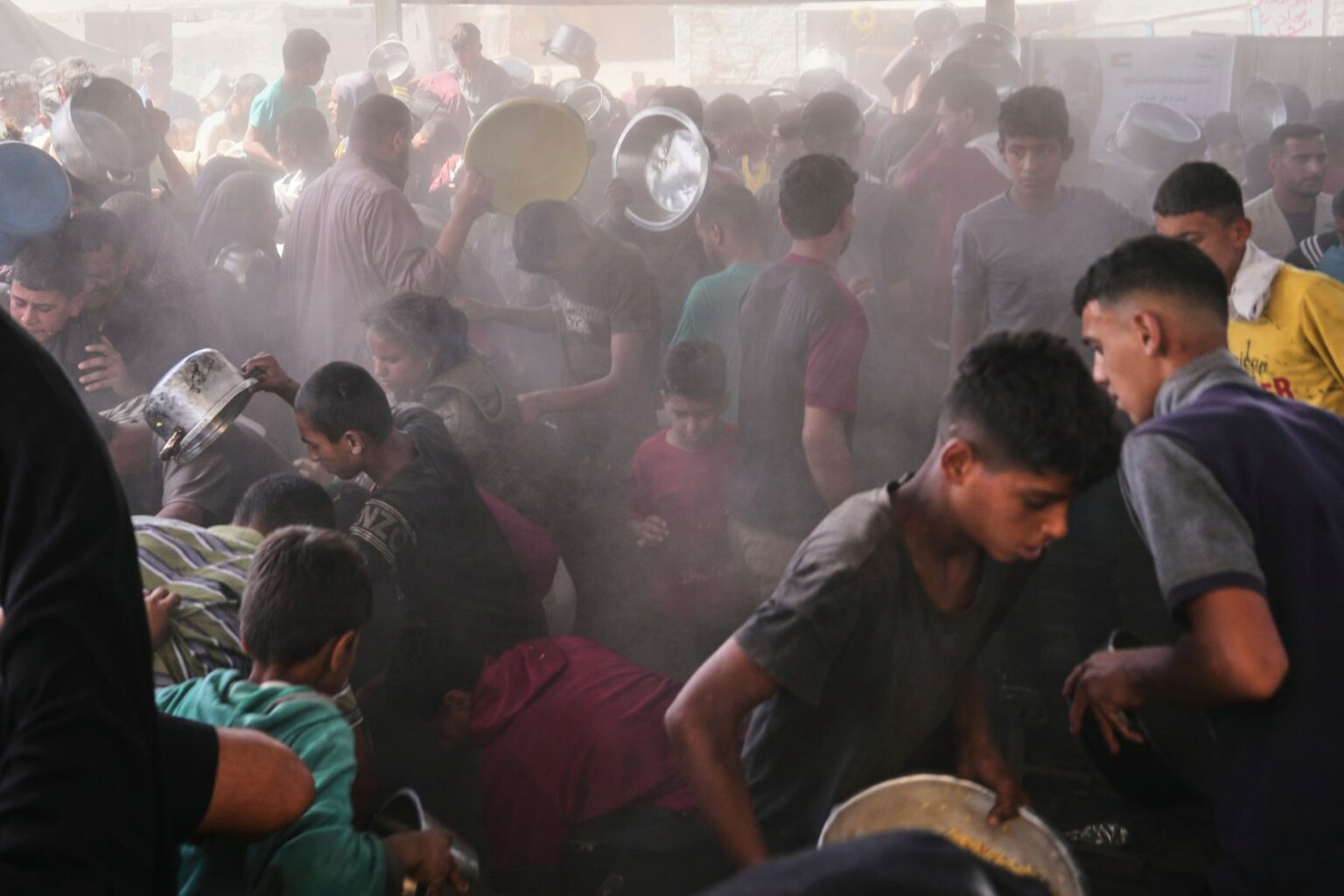 A photo of a dusty crowd of people holding pots and pans in the hopes of receiving food.