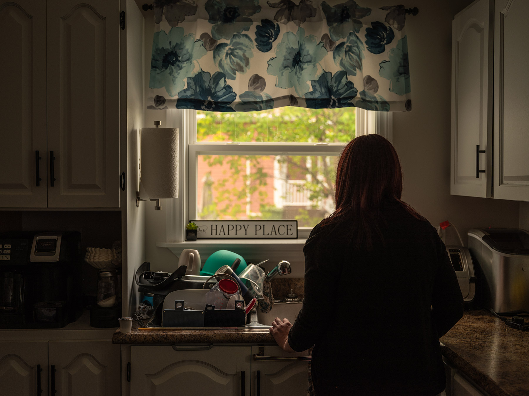 Photo of Jane Doe standing in her kitchen. She is facing away from the camera, looking out the window above the sink.
