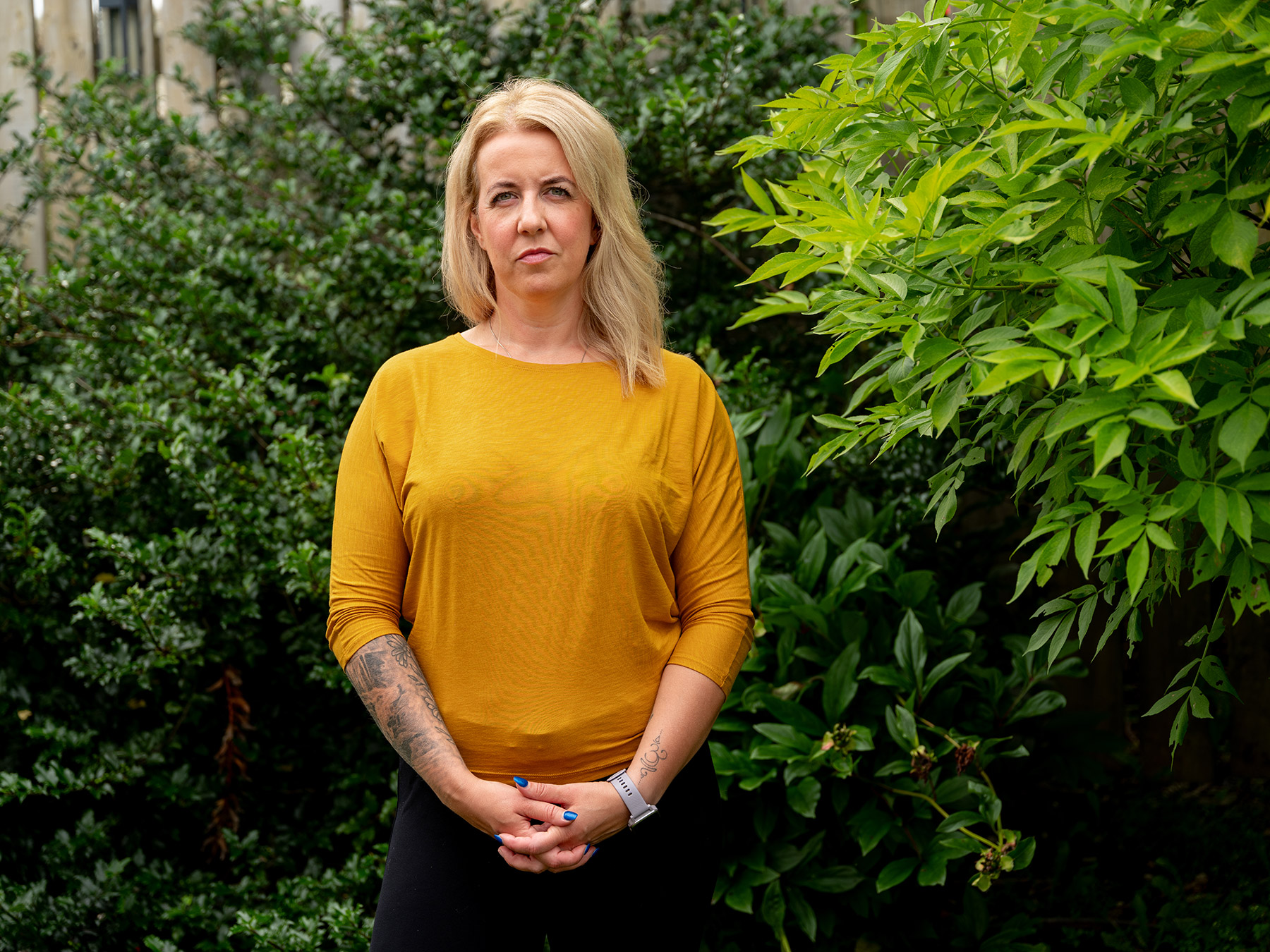 Photo of Kelsey Muise standing in front of green bushes outside of her home. She has shoulder-length blonde hair and is wearing a yellow sweater, hands clasped in front of her.