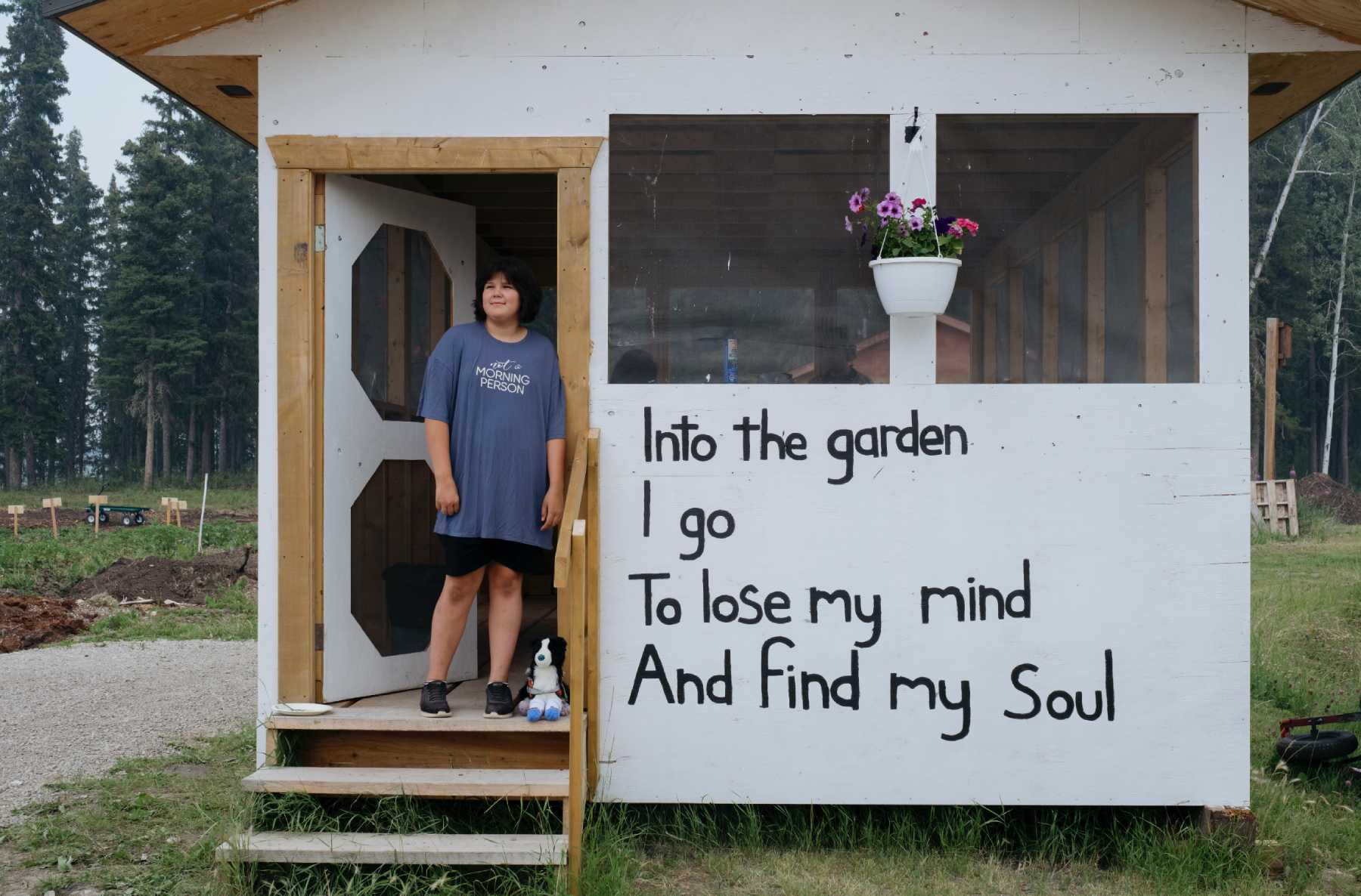 An image of a woman standing in the doorway of a white garden shed. There is a poem written on the barn: “I go to the garden To lose my mind And find my soul.”