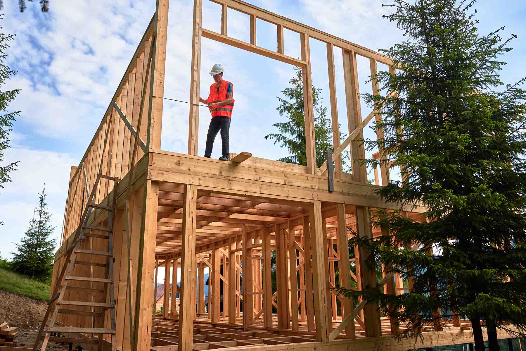 A man in an orange vest uses a tape measure while building a wooden frame house surrounded by trees.