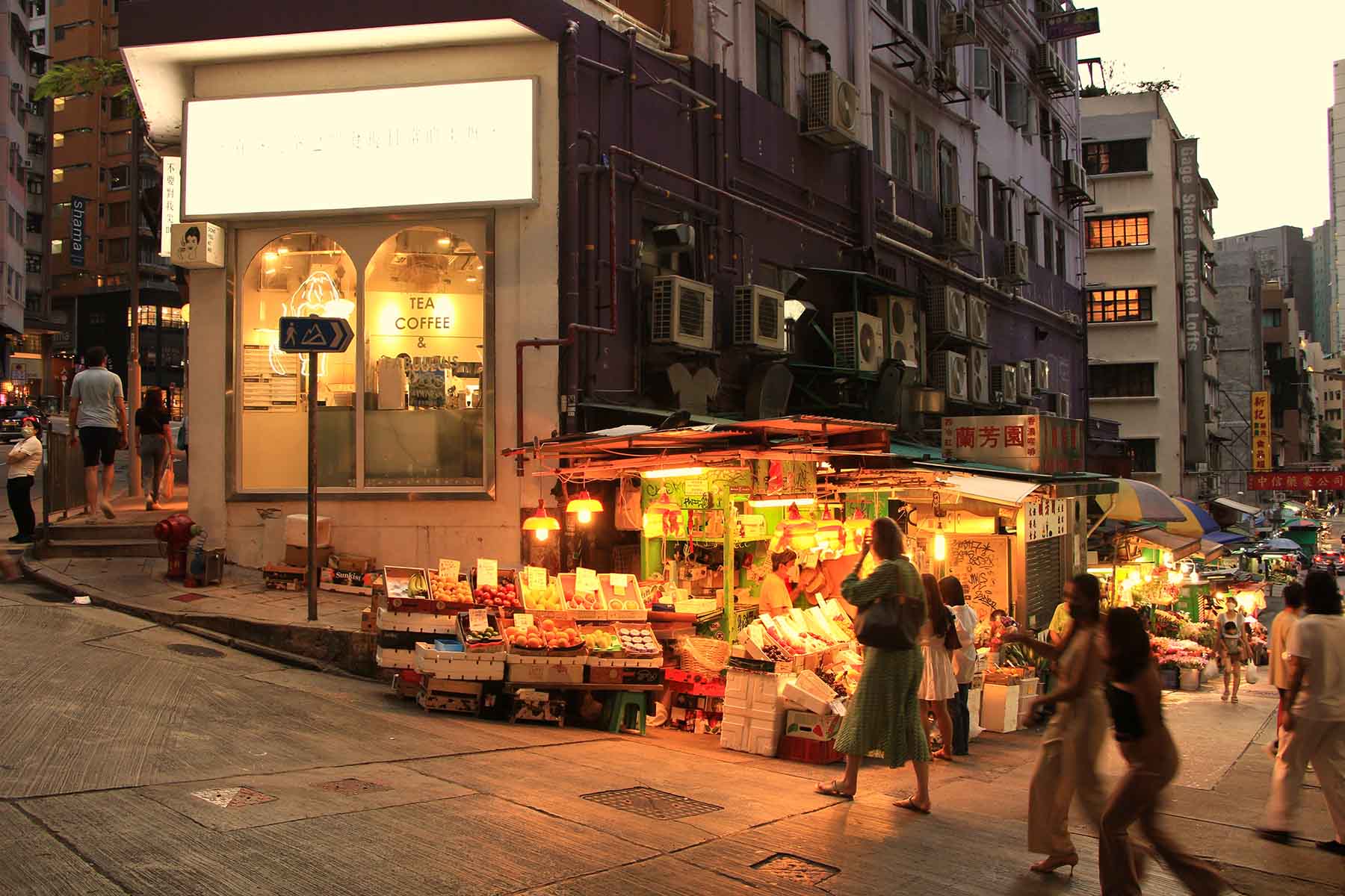 In the evening, people walk past a small fruit market illuminated by yellow light near a tea and coffee shop in Hong Kong.