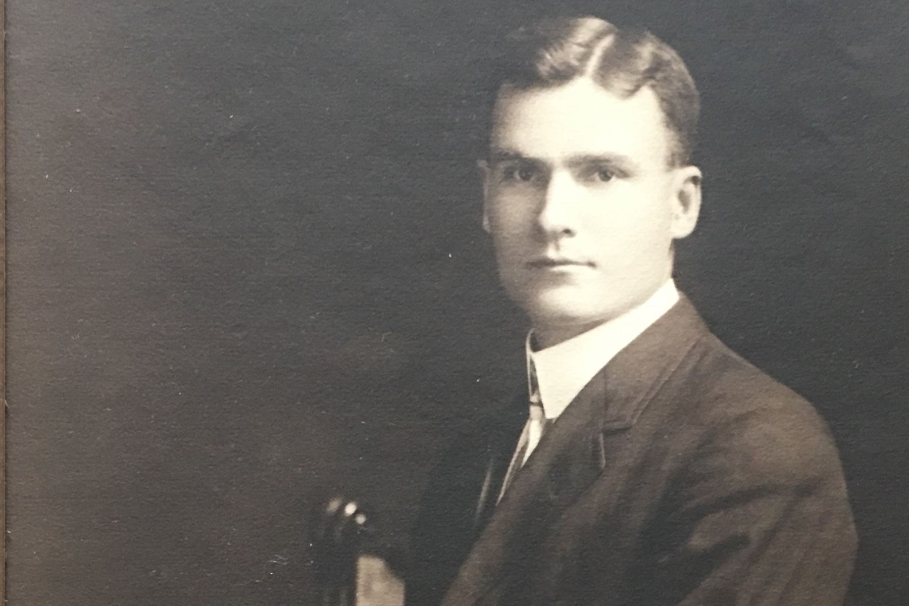 A sepia-tinged photo shows the portrait of a young man wearing a suit and looking at the camera while seated in a chair