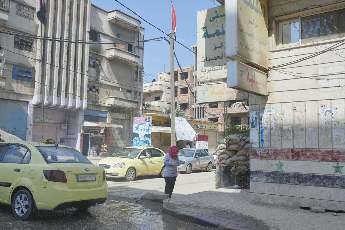 A woman stands on a street corner surrounded by cars and buildings.