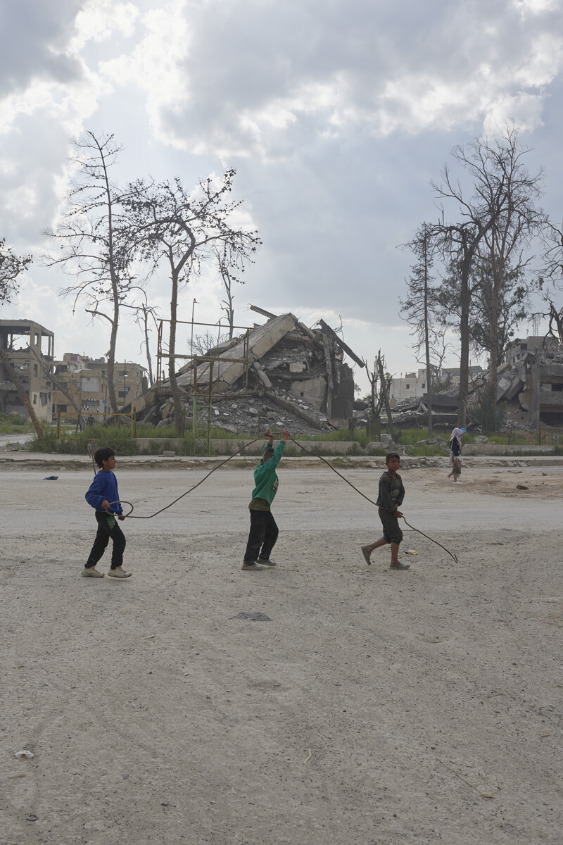 A line of three young children playing in a bare field in front of building rubble.