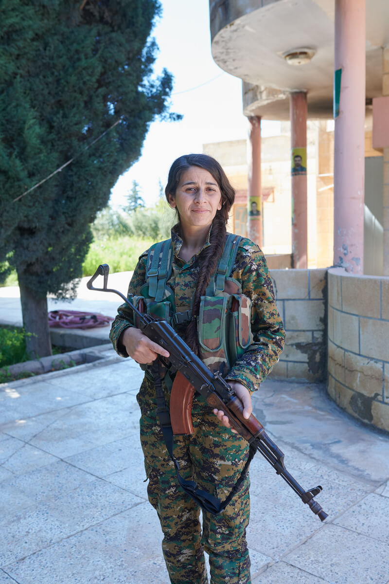 A woman wearing military fatigues and carrying a rifle faces the camera.