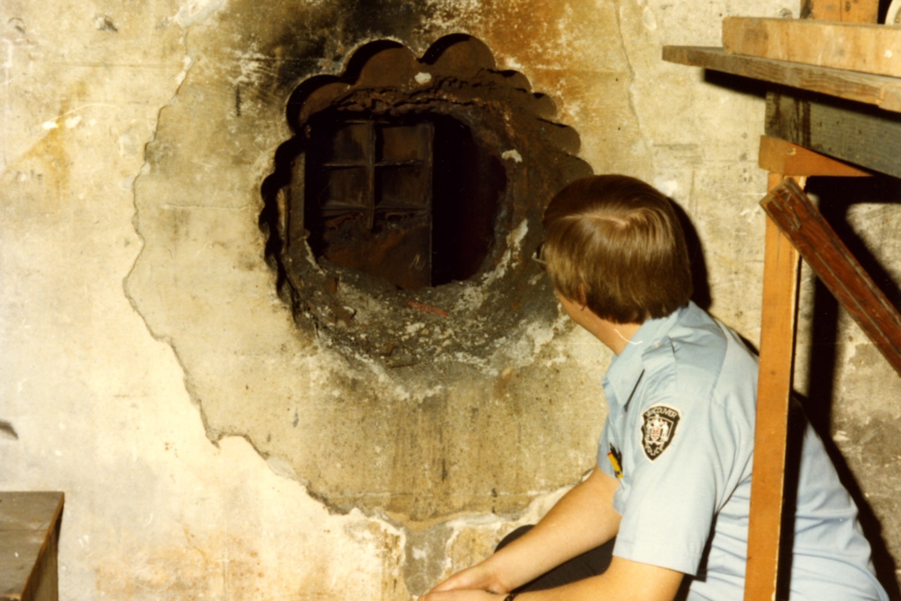 Photo of a man sitting next to a hole dug through a beige wall.