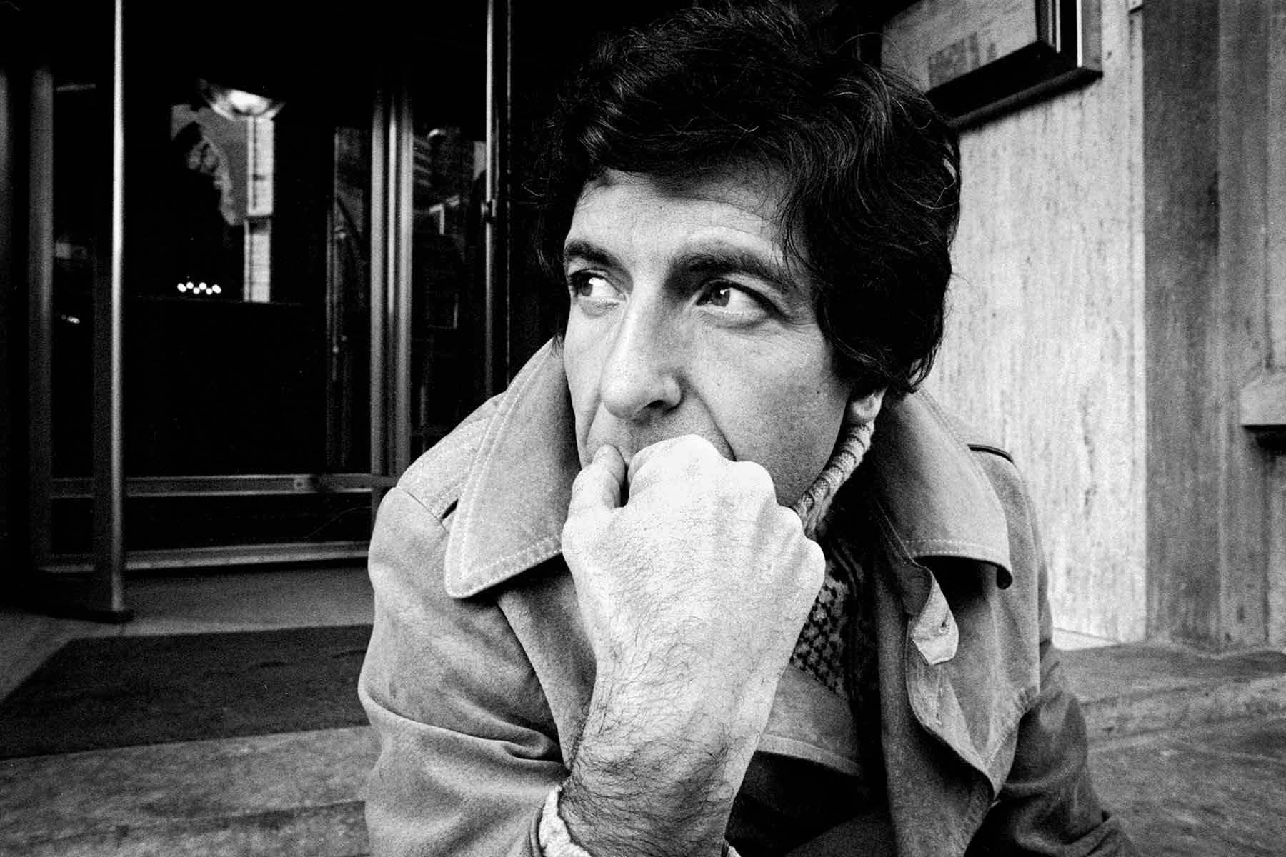 A black-and-white photo of Leonard Cohen sitting in front of a building with his hand obscuring his mouth.