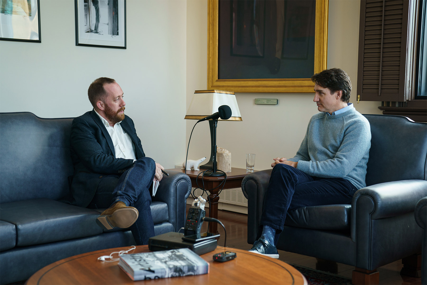 Journalist Justin Ling sits on a blue couch across from Justin Trudeau, seated in a blue arm chair with a microphone on the table between them