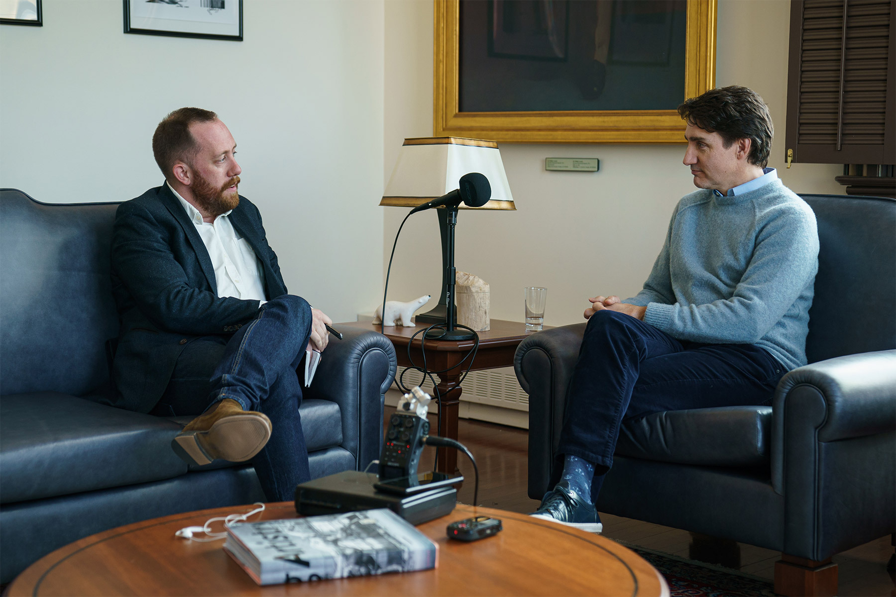 Journalist Justin Ling sits on a blue couch across from Justin Trudeau, seated in a blue arm chair with a microphone on the table between them.