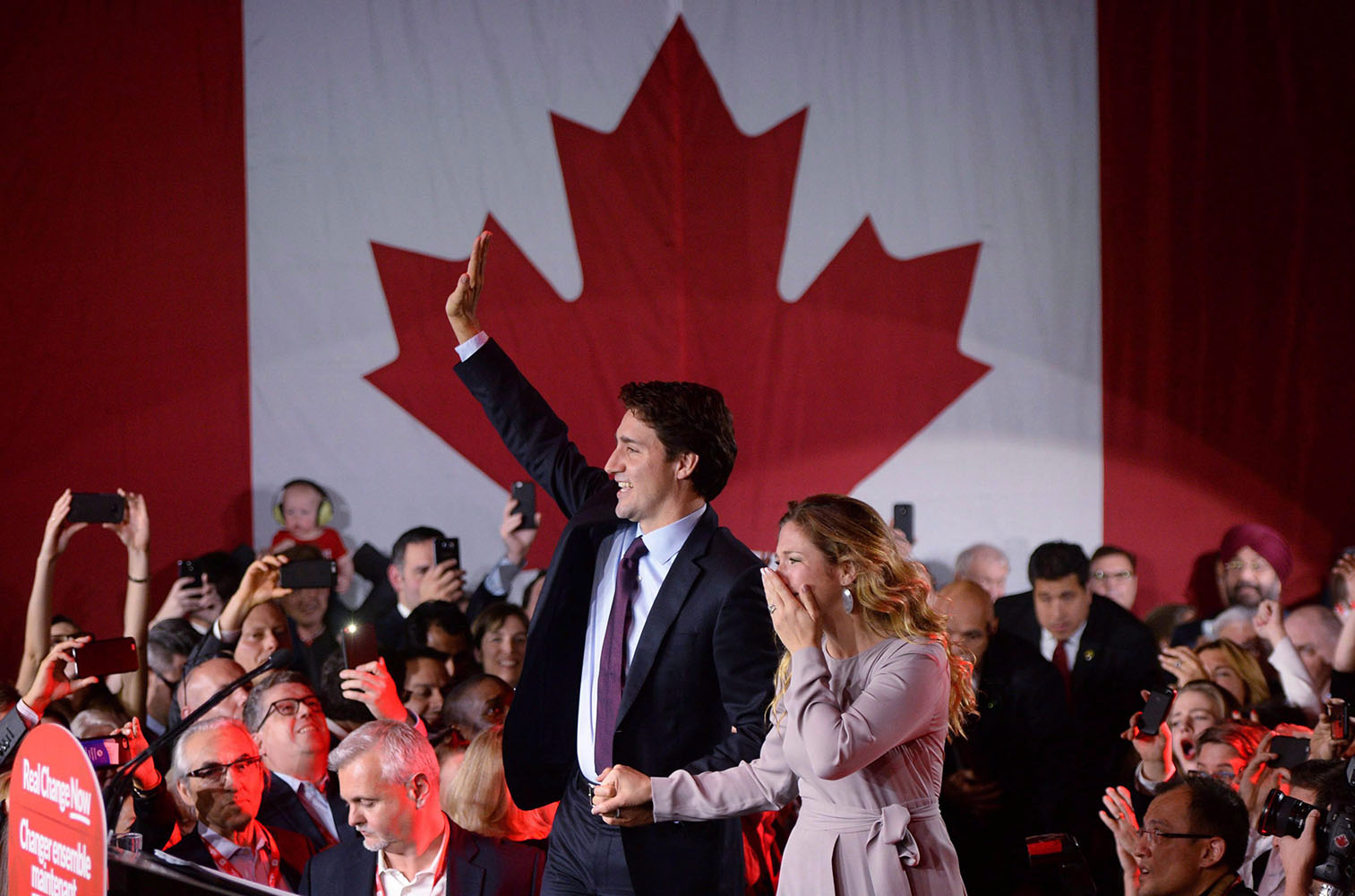 Justin Trudeau and Sophie Grégoire-Trudeau hold hands and wave to a crowd. In the background is a large Canadian flag.