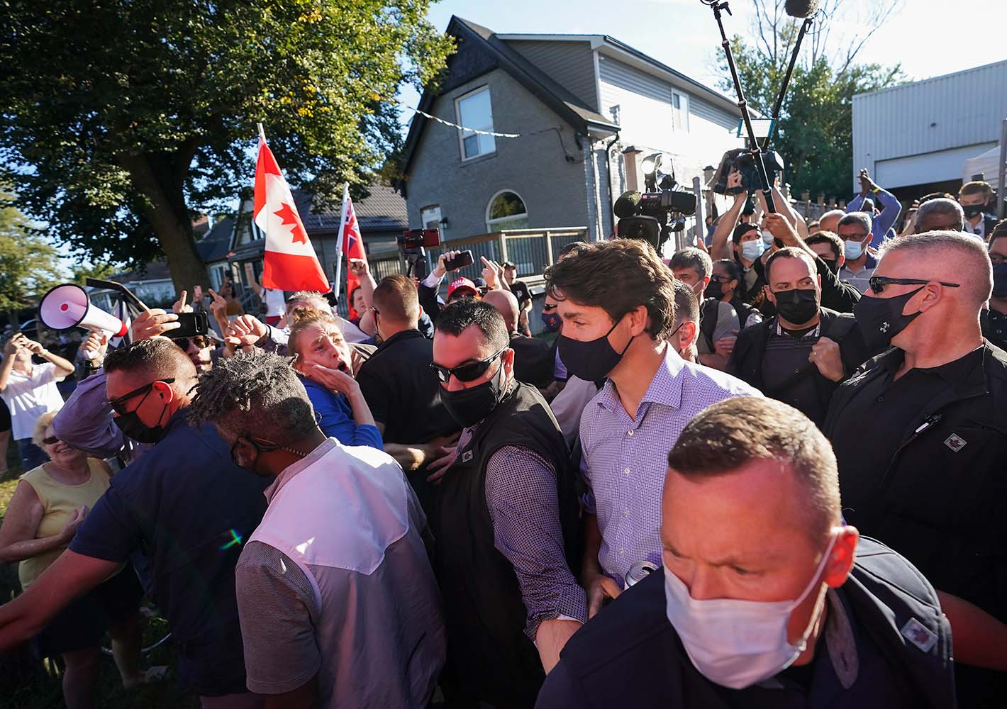 Justin Trudeau walks through a crowd of protesters, some yelling and holding Canadian flags, while surrounded by RCMP security personnel. They are wearing masks due to the COVID-19 pandemic.