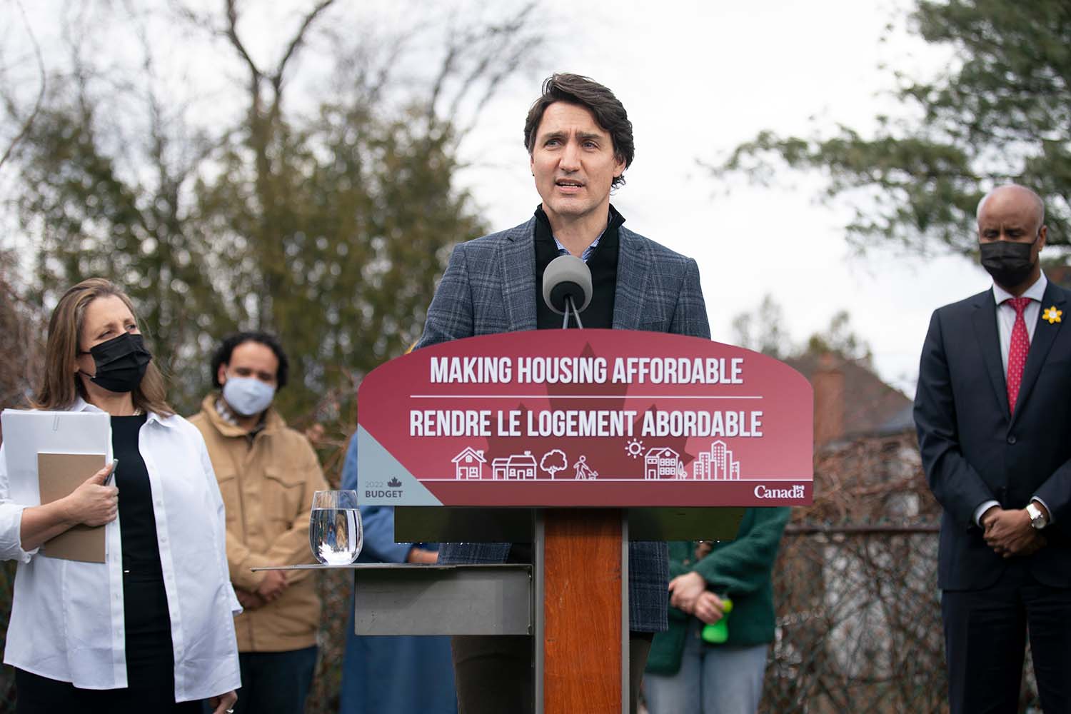 Justin Trudeau stands outside at a podium which says 'Making Housing Affordable / Rendre Le Logement Abordable.' Chrystia Freeland and several other staffers stand around him, wearing masks due to the COVID-19 pandemic.