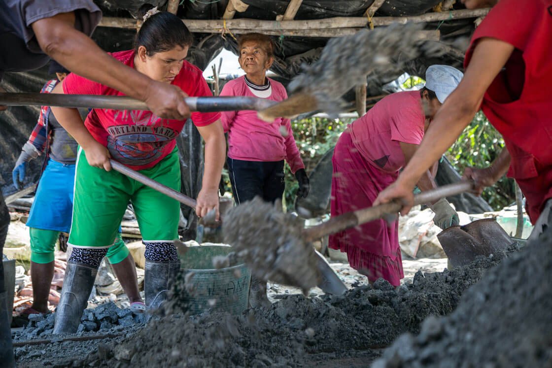 A group of women sift through buckets of mining ore with shovels.