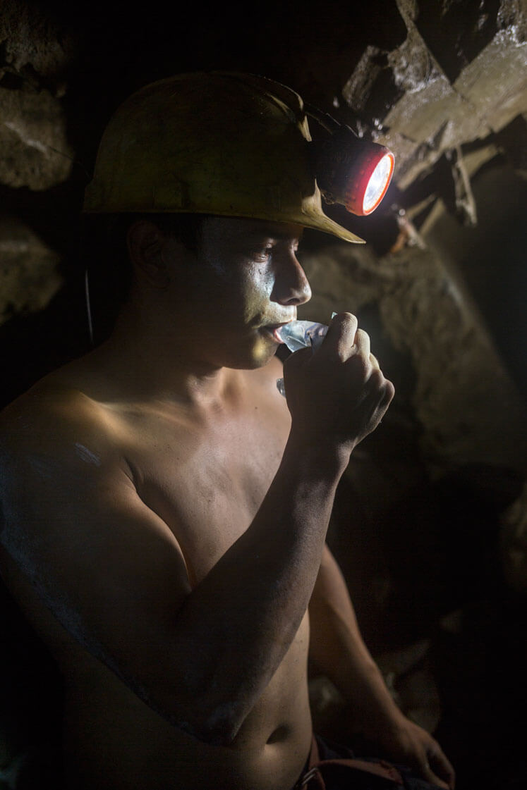 A miner wearing a hard hat and standing in a dark cave drinks water from a small plastic bag.