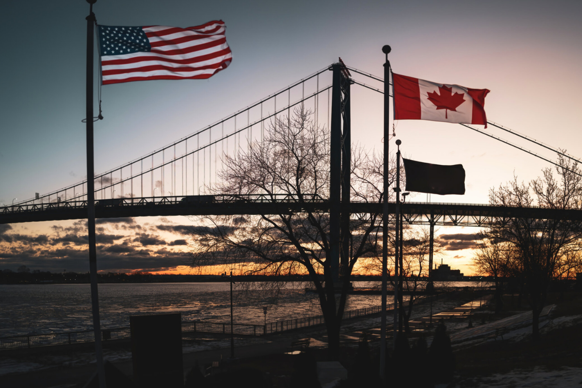 Photo
of a bridge at sunset. In the foreground, Canadian and American flags fly on flagpoles.