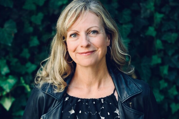 Photo of a woman with blonde hair smiling at the camera while standing in front of a leafy background.
