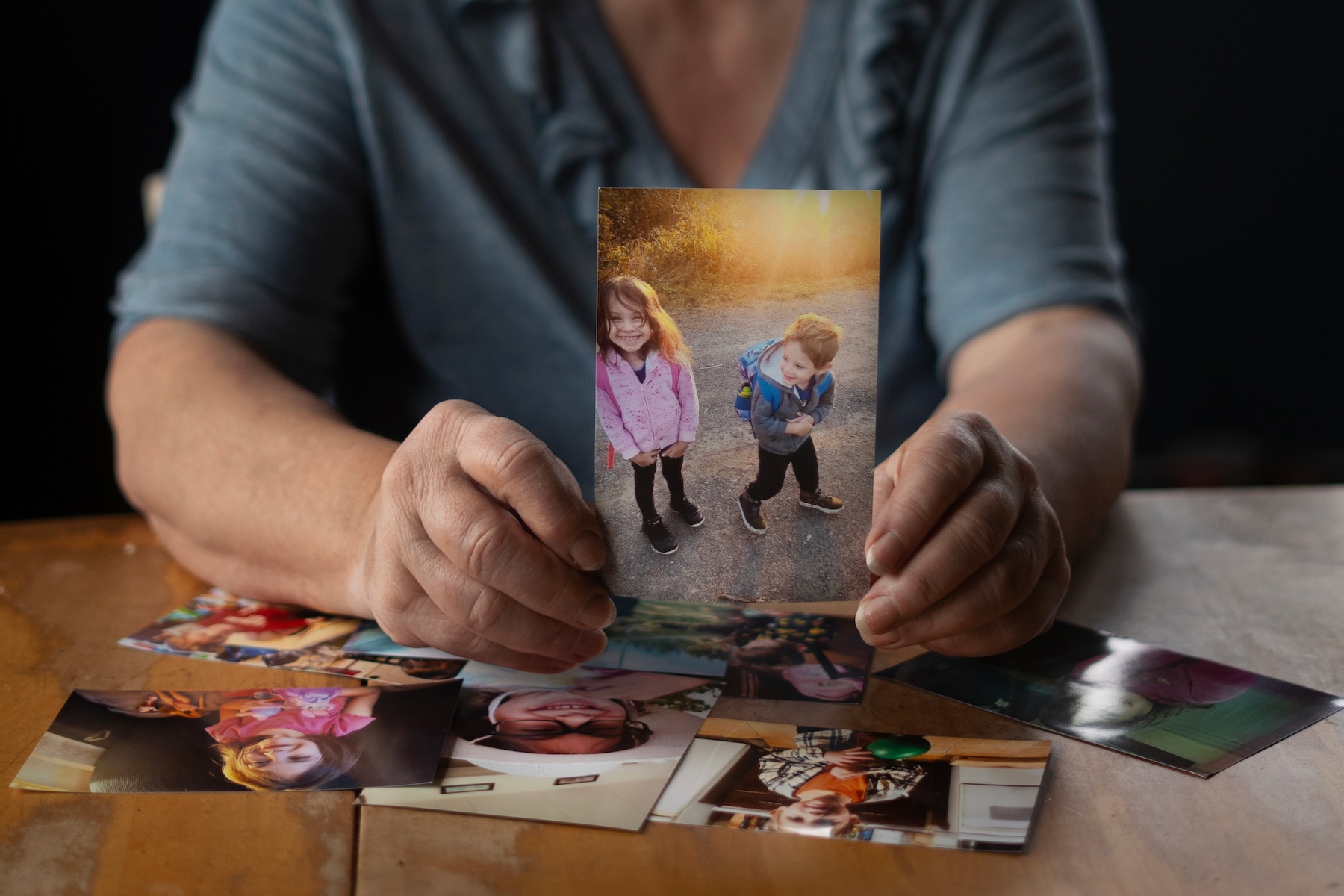 Photo of an elderly woman holding a printed picture of a young boy and girl in spring jackets. Other printed photos are scattered on the tabletop under her hands.
