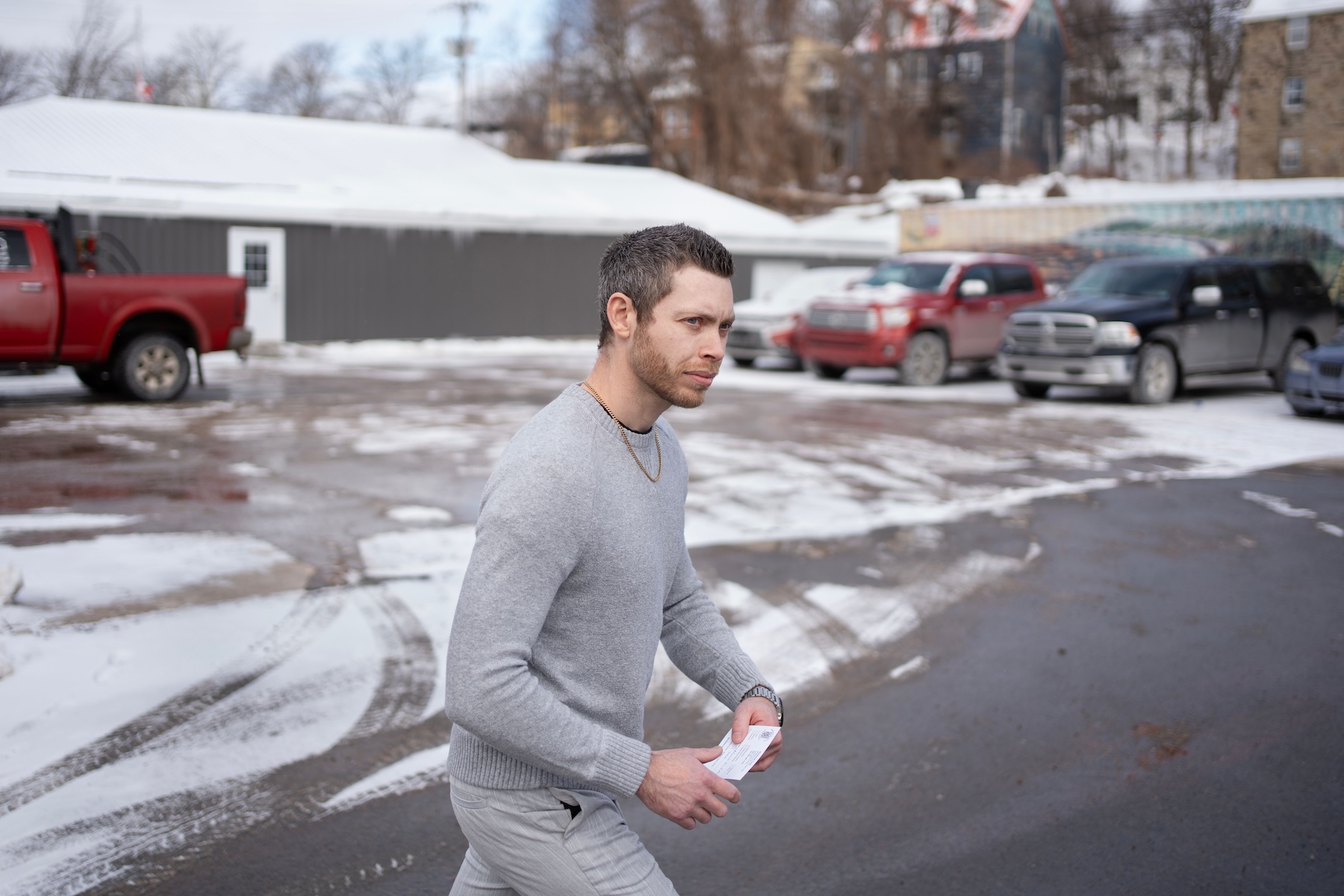 Photo of a man with short, brown hair wearing a grey shirt walking through a parking lot.