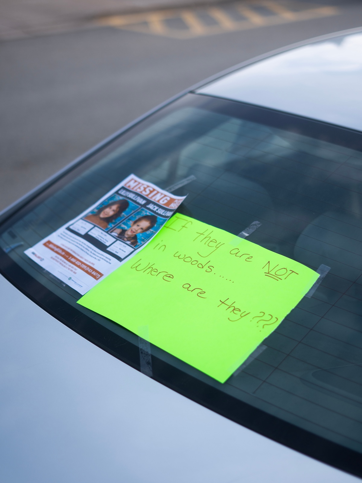 Photo of a missing poster depicting a young girl and boy and a sign on neon green paper which reads 'If they are not in the woods...where are they???' on a white car's windshield.