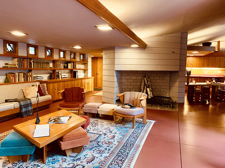 A photo of a living room in a Frank Lloyd Wright house, with slanted ceiling and a lot of wood. There are armchairs, a fireplace, and bookshelves on the walls.