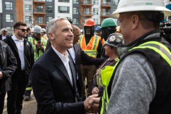 A photo of Mark Carney smiling and shking ther hand of a man in a construction helmet and safety vest. Behind them are other construction workers in hard hats and orange vests.
