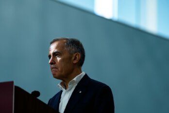 A photo of Prime Minister Mark Carney standing in front of a podium with a blue-grey backdrop behind him