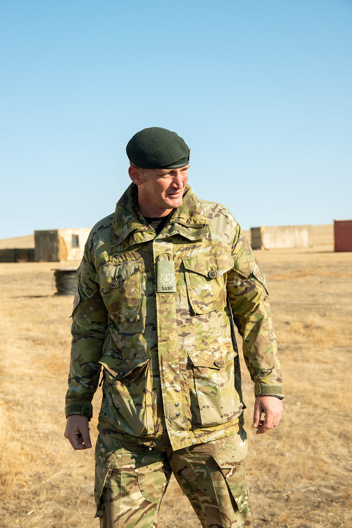 Photo of Terry Woods, wearing camo and a beret, standing in front of structures in a training village