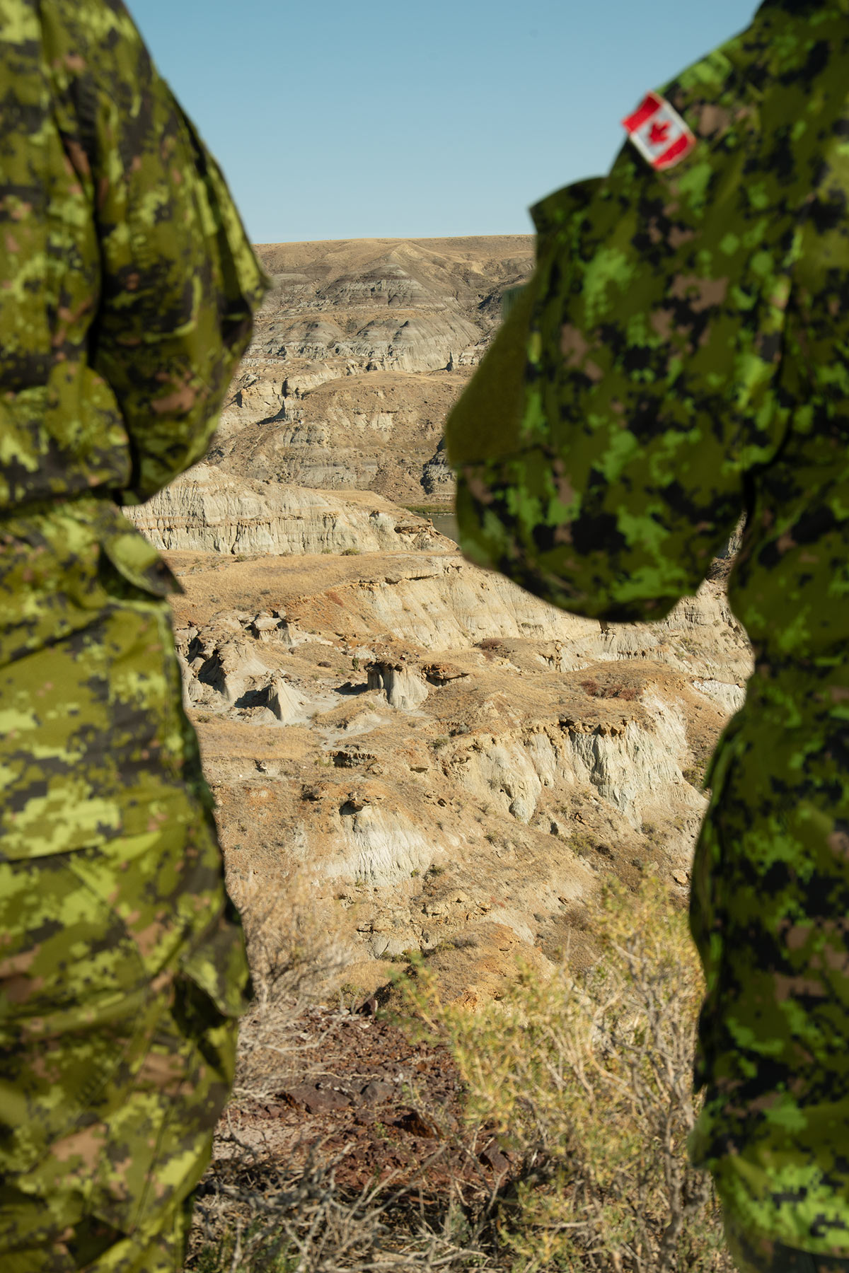 Photo of rocky beige landscape framed by the bodies of two military personnel wearing green camo. A small Canadian flag patch is seen on the arm of the person on the right.