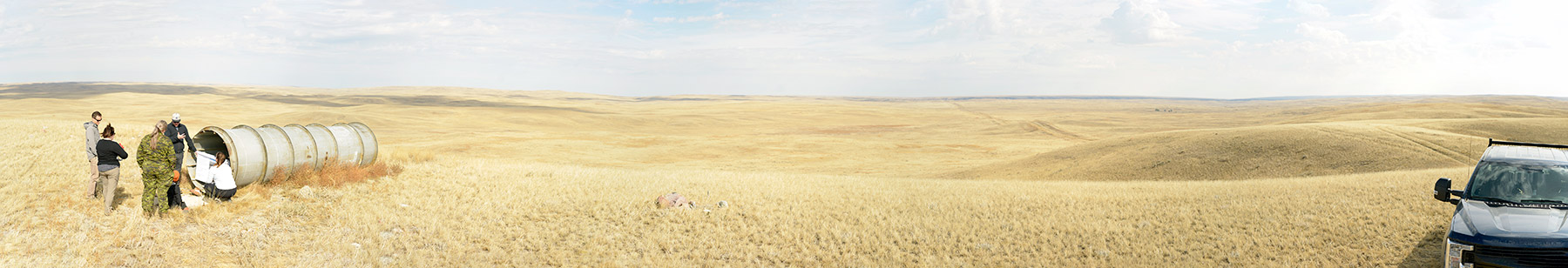 Panoramic photo of a rolling landscape with yellow fields under a blue sky. Military personnel in camo and researchers are seen by equipment on the left and a truck is parked on the far right