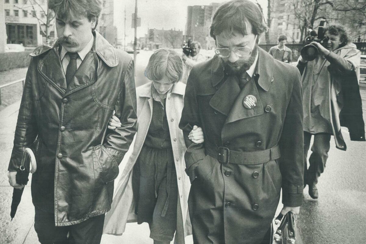 A black-and-white photo of a petite woman, head down, walking with her hands of the arms two taller man. Behind them, a grimacing man films them with a video camera on his shoulder