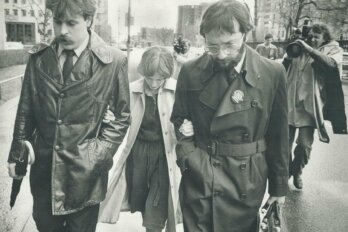 A black-and-white photo of a petite woman, head down, walking with her hands of the arms two taller man. Behind them, a grimacing man films them with a video camera on his shoulder