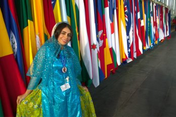 Ghanimat Azhdari, a young woman in a blue dress, poses and smiles against a backdrop of nations' flags.