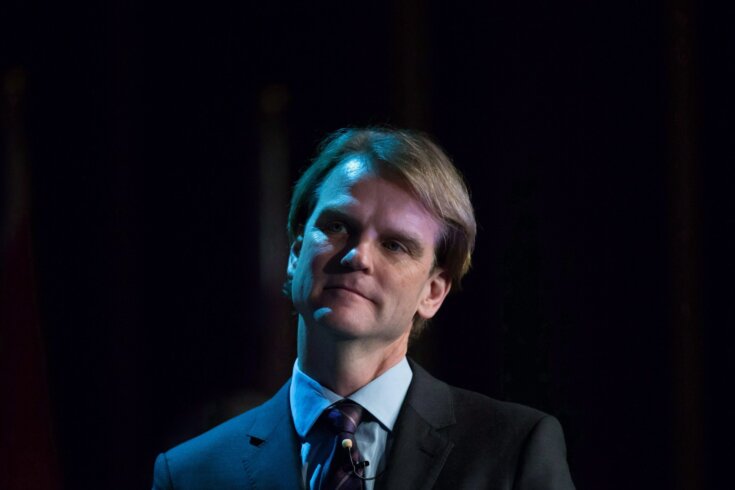 Headshot photo of a man with short brown hair in a suit against a black stage background.