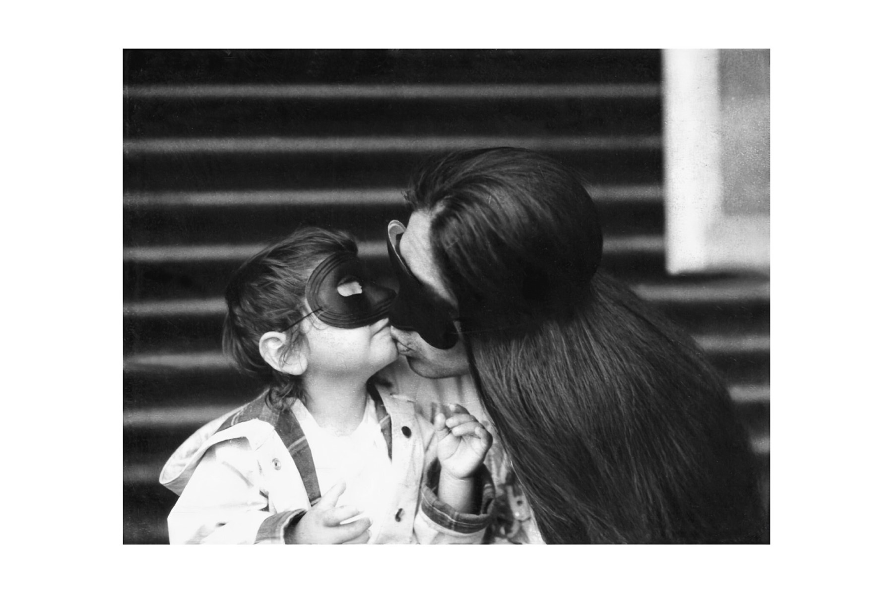 Black and white photo of a person with long hair kissing a young boy. Both are wearing black costume masks over their eyes.