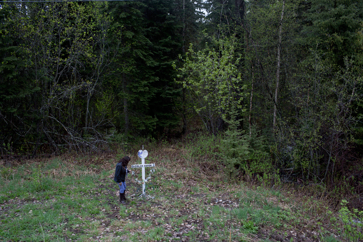 A woman stands by a small memorial, a cross marked with the name Aielah Auger. The memorial is in the grass at the side of a highway, immediately before the forest begins.